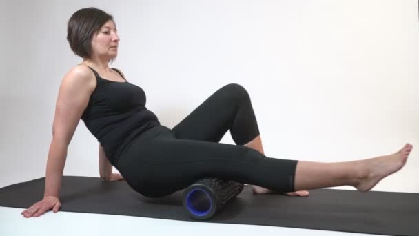 Una mujer de mediana edad sobre una colchoneta de gimnasia con rodillo miofascial hace un ejercicio en sus caderas sobre un fondo blanco . — Vídeos de Stock