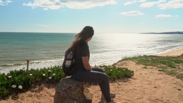Ein erfolgreiches, glückliches junges Mädchen setzte sich auf eine Klippe in der Nähe des Ozeans, müde von einem Spaziergang, um sich auf einem Stein auszuruhen. Portugal. Vilamoura. — Stockvideo