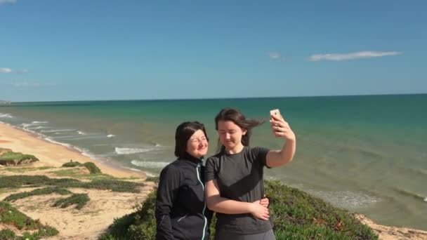 Başarılı, orta yaşlı bir kadın ve genç bir kız okyanusun yakınındaki bir uçurumun kenarında telefonla selfie çekiyorlar. Portekiz. Vilamoura. Anne ve kızı.. — Stok video