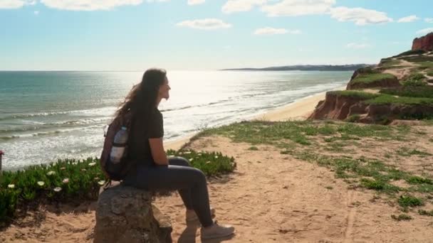 Uma jovem bem-sucedida e feliz, em um penhasco perto do oceano, cansada de um passeio, sentou-se para descansar em uma pedra. Portugal. Vilamoura . — Vídeo de Stock