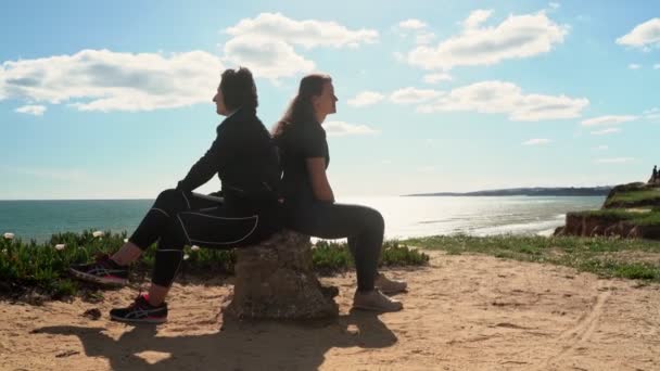 Başarılı, orta yaşlı bir kadın, genç bir kızla birlikte okyanusun kenarındaki bir uçurumda yorgun bir şekilde bir taşın üzerine oturmuş. Portekiz. Vilamoura. Anne ve kızı.. — Stok video