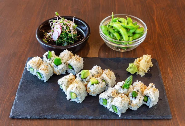 Japansk sushimat med avokado, ris, havets delikatesser på en svart sten, på en brun bakgrund. Plattor med sallad och gröna bönor. — Stockfoto