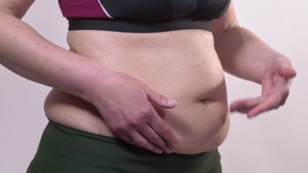 Eine Frau mittleren Alters mit schlaffer Haut schüttelt vor weißem Hintergrund einen dicken Bauch. Seitenplan. — Stockvideo