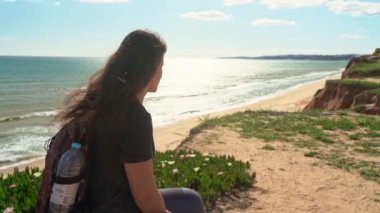 Başarılı, mutlu genç bir kız, okyanusun yakınındaki bir uçurumda, yürümekten yorulmuş, bir taşın üzerine oturmuş. Portekiz. Vilamoura.