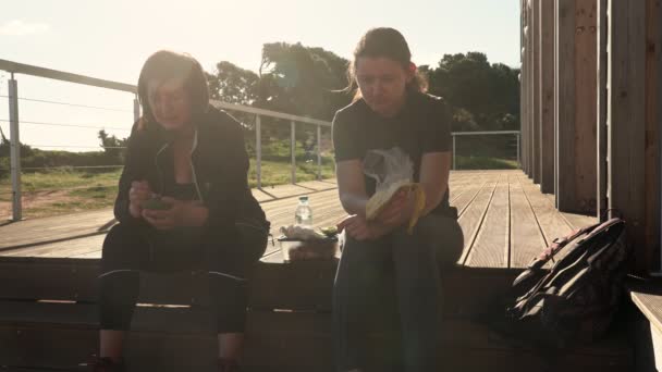 Een succesvolle, gelukkige vrouw van middelbare leeftijd en een jong meisje, moe van een sportieve wandeling, gingen zitten om wat te eten op een houten trap. — Stockvideo