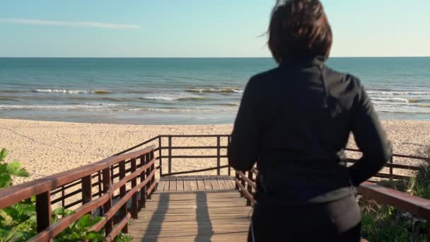 Mulher de meia-idade feliz corre em uma passarela de madeira para o oceano. Movimento lento . — Vídeo de Stock