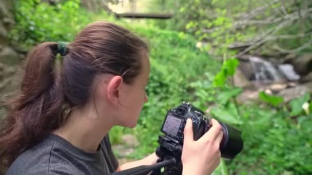 Egy fiatal lány, egy fotós, képeket készít a gyönyörű természetről egy erdős területen, ahol patak folyik.. — Stock videók
