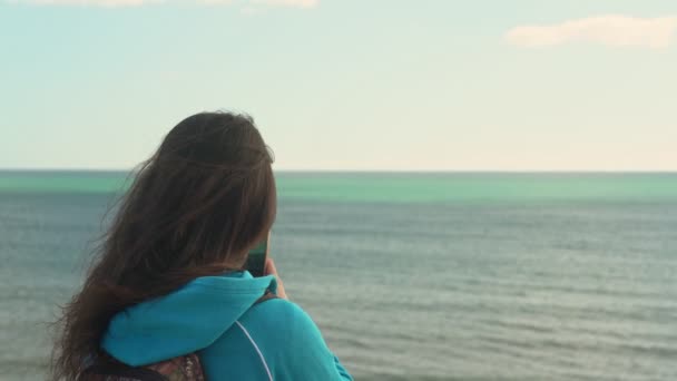 Jong meisje toerist op het strand door de oceaan met een telefoon in zijn hand zal foto 's van het landschap om hem heen te nemen. — Stockvideo
