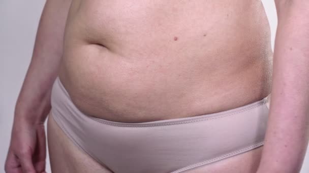 Žena středního věku s povislou pletí definuje své velké břicho měřicí páskou se zatáčkami na bílém pozadí. Vedlejší plán. — Stock video