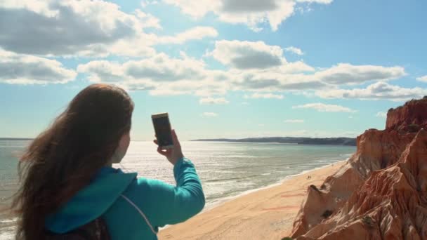 Молода дівчина турист на пляжі біля океану з телефоном в руці зробить фотографії ландшафту навколо нього . — стокове відео