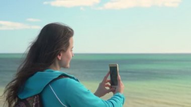 Okyanus kıyısındaki genç bir turist elinde telefonla etrafındaki manzaranın resimlerini çekecek..