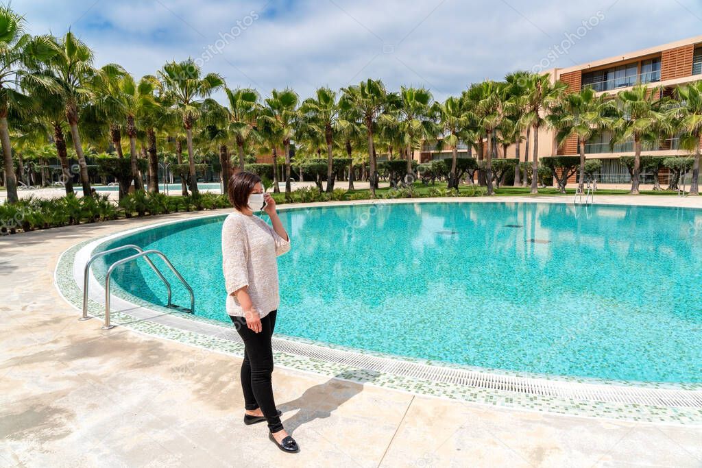 A middle-aged woman in a protective mask against the virus, coronavirus, and allergies is talking on the phone near the pool in the hotel.