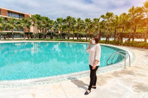 Una mujer de mediana edad, en un día soleado, con una máscara protectora contra el virus, coronavirus, alergias, hablando por teléfono cerca de la piscina en el hotel . —  Fotos de Stock