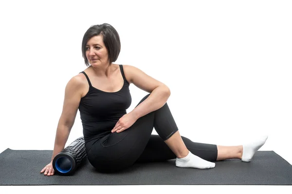 Una donna di mezza età, su un tappeto da ginnastica con rullo miofasciale, sta facendo un esercizio di torsione su uno sfondo bianco . — Foto Stock
