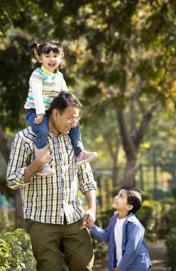 Babası kızı omuzlarında taşıyor ve parkta oğlunun elini tutuyor.