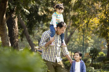 Babası kızı omuzlarında taşıyor ve parkta oğlunun elini tutuyor.