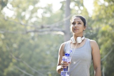 Sporcu kadın parkta su içiyor ve müzik dinliyor.