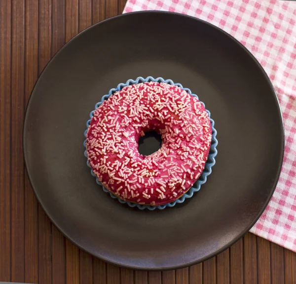 Ярко розовый пончик на черной керамической тарелке на коричневом деревянном пляже — стоковое фото
