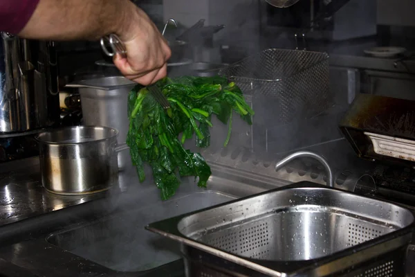 Повар достает из соуса отварной зеленый горячий шпинат. — стоковое фото