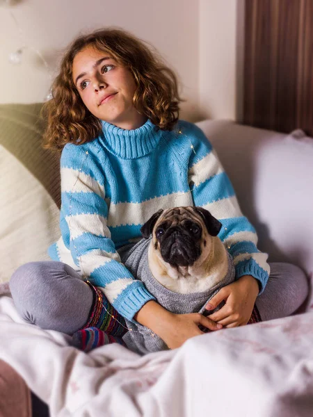 Красивая девочка-подросток обнимает мопса с любовью. Кудрявая девушка в вязаном свитере держит мопса . — стоковое фото