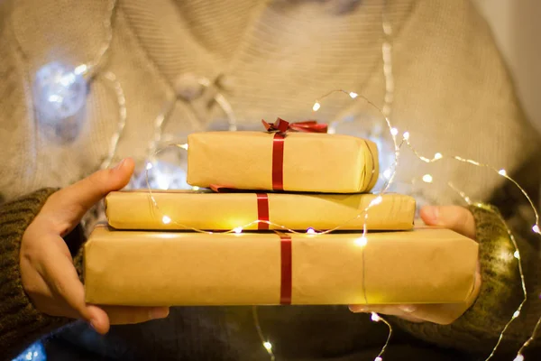 Gift boxes with red ribbon in the hands. Hands hold gifts in craft paper with luminous garland — ストック写真