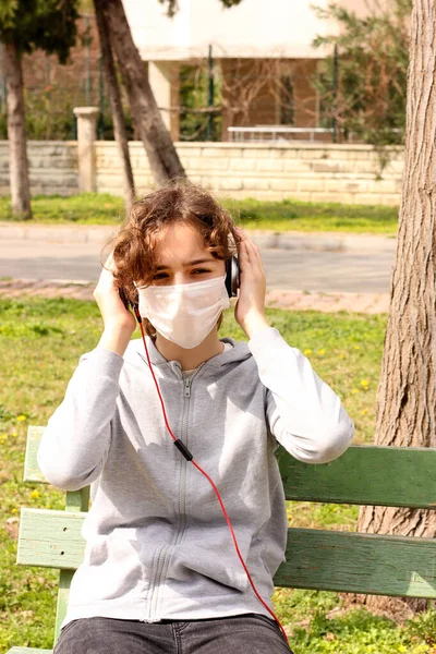 Young teenager girl with a medical mask is listening to music with headphones in park. Home education, quarantine and isolation during coronavirus