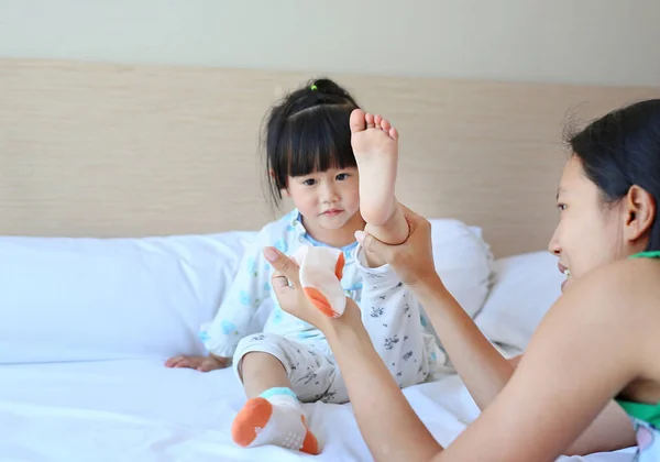 Mother trying to put a socks for her daughter on the bed.