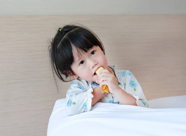 Ładna dziewczynka w piżamie jedzenie banana na łóżku w nocy. — Zdjęcie stockowe