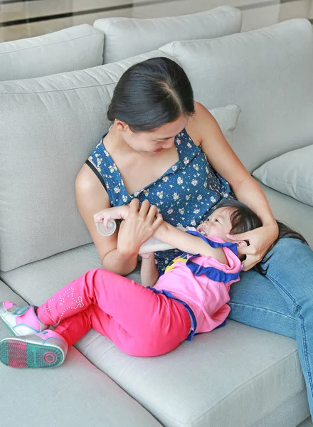 Ibu memeluknya usia anak perempuan Asia yang lucu sekitar satu tahun dan sembilan bulan minum dari botol di sofa . — Stok Foto