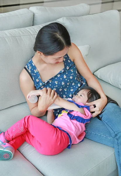 Mother hug her Cute asian child girl age about one year old and nine month drinking from a bottle on the sofa. — Stock Photo, Image