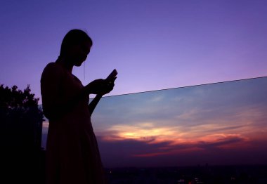 Binanın çatı üzerinde gün batımında smartphone kullanan kadın silüeti