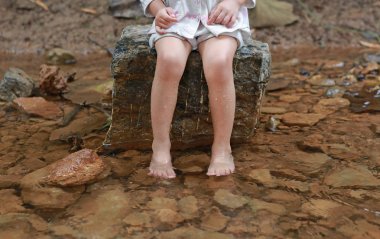 Legs of little child girl sitting on stone and playing water, countryside thailand. clipart