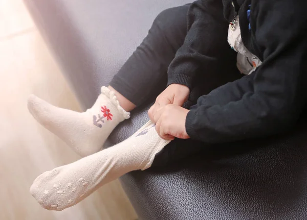 Niña tratando de poner un calcetines en el sofá marrón . —  Fotos de Stock