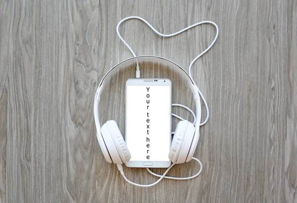 Headphones and smartphone with word "Your text here" on white screen against wooden background — Stock Photo, Image