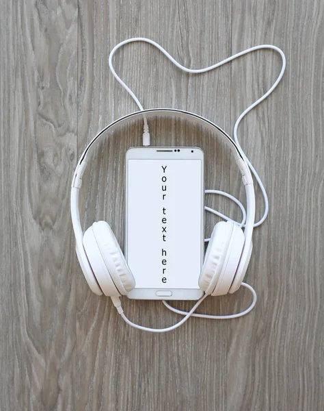 Headphones and smartphone with word "Your text here" on white screen against wooden background — Stock Photo, Image
