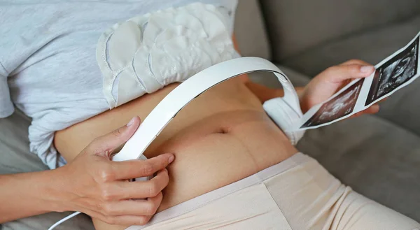 Portrait of pregnant woman sitting on sofa at home holding ultrasound film and listening music in headphones.