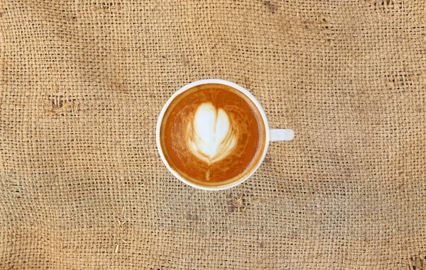 Vista dall'alto di un caffè con motivo a cuore in una tazza bianca su sfondo sacco, arte del latte — Foto Stock