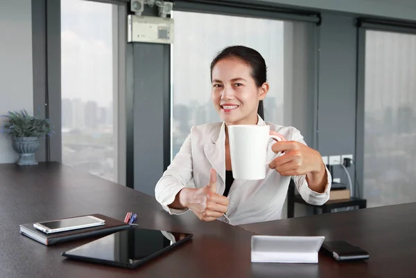 Young businesswoman sitting at the table on workplace in office show thumb up with holding a coffee cup and smiling — Stock Photo, Image