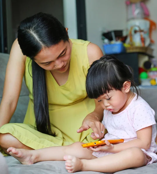 Ibu dan bayi perempuan asia yang lucu bermain smartphone dan berbaring di sofa abu-abu di ruang tamu — Stok Foto