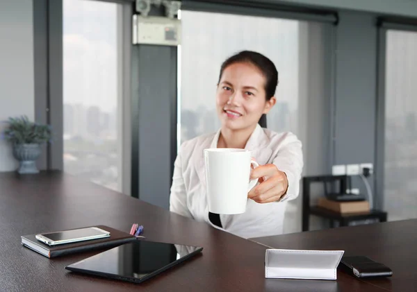 Young businesswoman sitting at the table on workplace in office offers a white cup of coffee, Focus at the coffee cup — Stock Photo, Image
