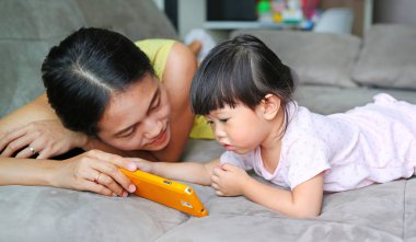 Anne ve sevimli Asya bebek kız yaşı hakkında bir yaşında ve sekiz ay bir akıllı telefon çalmaya ve gri kanepe oturma odasında döşeme