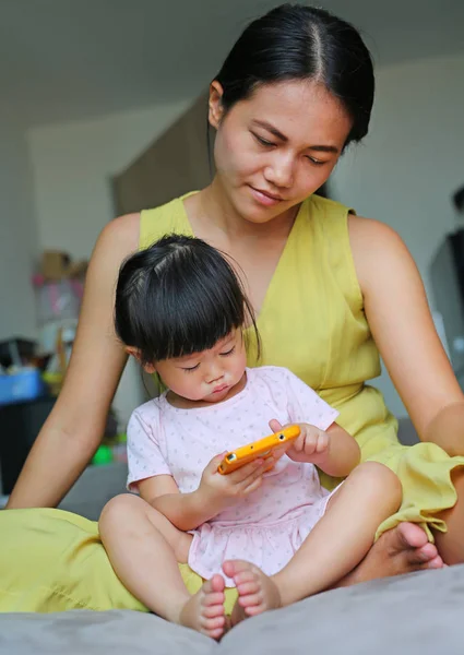 Madre y linda niña asiática edad alrededor de un año y ocho meses jugando un teléfono inteligente y acostado en un sofá gris en la sala de estar —  Fotos de Stock