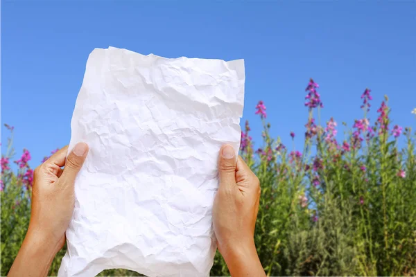 Frauenhände halten quadratisches zerknülltes Papier vor verschwommenem Hintergrund der Natur und blauem Himmel. — Stockfoto