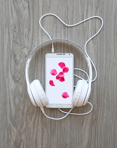 Sluchátka a smartphone s lístky růžové růže na bílé plátno dřevěné pozadí — Stock fotografie