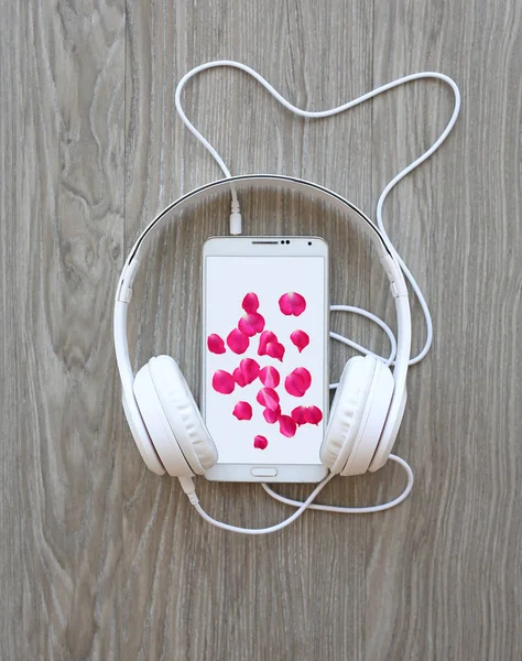 Écouteurs et smartphone avec pétales de rose sur écran blanc sur fond bois — Photo