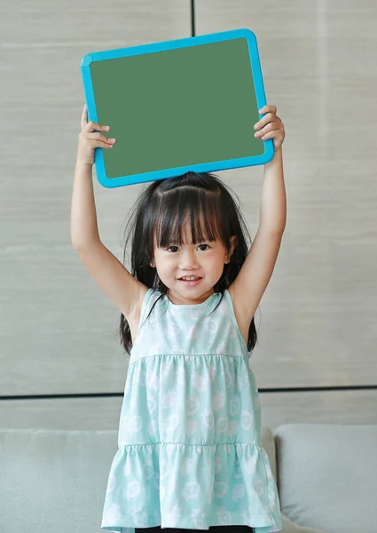 Cute little child girl holding empty green blackboard standing on fabric sofa in library room. Education concept. — Stock Photo, Image