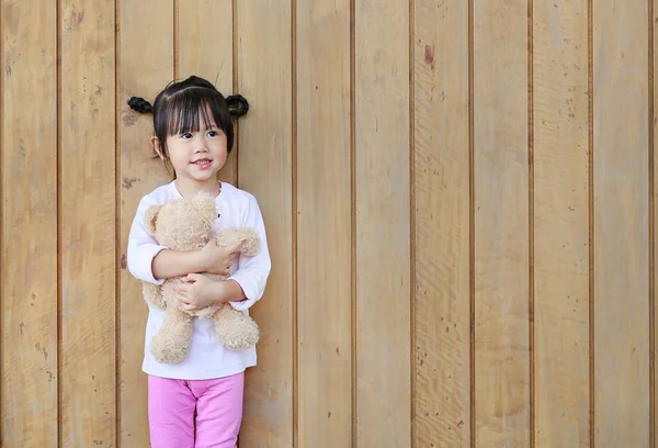 Retrato de bonitinho menina stand e abraçando ursinho de pelúcia contra a parede da prancha de madeira — Fotografia de Stock