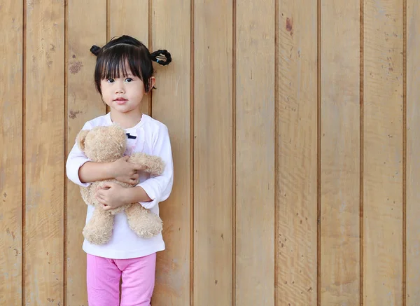 Retrato de bonitinho menina stand e abraçando ursinho de pelúcia contra a parede da prancha de madeira — Fotografia de Stock