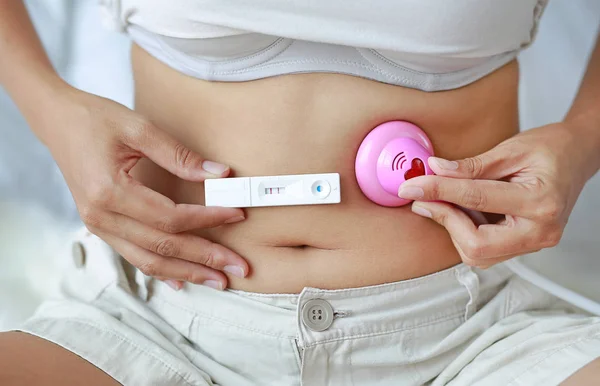 Woman holding positive pregnancy test at her belly with toy stethoscope.