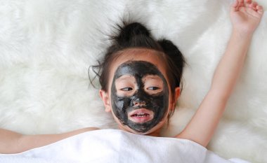 Kid girl in coal peeling face mask, beauty concept. clipart